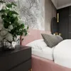 A close-up view of a pink upholstered bed with white and gray pillows, next to a dark bedside table with a vase of white flowers. Náhľad obrázku produktu