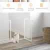 A white metal pet gate is installed in a doorway, with a small white dog sitting in front of it. The background shows a wall and floor, with a wicker basket in the corner. Náhľad obrázku produktu