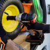 A person is using a WORX impact wrench to tighten a bolt on a yellow wheel, possibly of a wheelbarrow or garden cart. The scene appears to be in the back of a vehicle or storage area, with other items like an orange bucket and green straps visible. Podgląd zdjęcia produktu