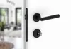 A close-up view of a black door handle and lock on a white door, with a blurred interior scene visible through the doorway. Produktbild-Vorschau