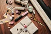 A collection of gift-wrapping supplies, including a BOSCH Gluey hot glue gun, scissors, ribbon, pom-poms, and gift tags, arranged on a wooden surface. Produktbild-Vorschau