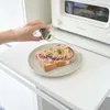 A hand is sprinkling herbs onto a piece of toast with cheese, ham, and onions, placed on a plate next to a white toaster oven. Podgląd zdjęcia produktu