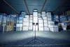 A warehouse interior with stacks of products and a tripod with two LED lights. Produktbild-Vorschau
