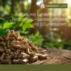 A pile of wood pellets in front of a blurred forest background with sunlight filtering through the trees. Produktbild-Vorschau