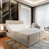 A bedroom scene featuring a large box spring bed with a beige upholstered headboard and base, set against a modern, textured wall. Two armchairs are visible in the background, along with sheer curtains. Produktbild-Vorschau
