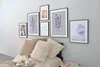 A collection of framed art prints displayed on a wall above a bed with several decorative pillows. Produktbild-Vorschau