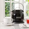 A black KitchenAid electric kettle is shown next to a white teacup on a saucer, with a glass of red juice and a white napkin with cutlery in the foreground. The scene is set in front of a window with greenery visible outside. Produktbild-Vorschau