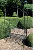 A black metal garden bench with an arched top, set in a garden with manicured bushes and trees. Produktbild-Vorschau