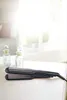 A hair straightener is placed on a white surface, with two decorative bottles in the background. Podgląd zdjęcia produktu
