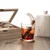 A tea infuser in a glass mug with tea, a tea box, and a teapot in the background. Produktbild-Vorschau
