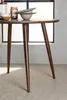 A close-up view of a round wooden table with three legs, set against a window with a light gray wall. Produktbild-Vorschau