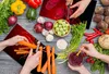 A top-down view of hands preparing vegetables on a wooden table. Anteprima dell’immagine prodotto