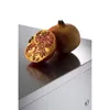 A cut pomegranate and a whole pomegranate are placed on a reflective surface, likely a kitchen counter or appliance. Anteprima dell’immagine prodotto
