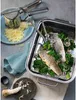 A prepared meal of fish and greens in a steamer basket, with cooking utensils and ingredients nearby. Náhľad obrázku produktu