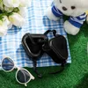 A teddy bear wearing a blue and white striped shirt sits on a green surface next to a blue and white checkered blanket. A pair of white sunglasses and black ear protection for dogs are also visible. Produktbild-Vorschau