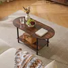 A dark brown oval coffee table with two shelves, placed on a light gray rug in a living room setting. The table has a vase with flowers, a bowl of fruit, a book, and a woven basket on it. Náhled obrázku produktu