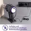 A cordless vacuum cleaner is charging on its stand. In the background, an electrical outlet is visible with the charging adapter plugged in. There is text and an icon at the bottom of the image. Produktbild-Vorschau
