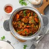 A close-up overhead view of a steaming pot of stew with carrots and mushrooms, garnished with parsley. A small bowl of spices and a wooden cutting board are visible in the background. Voir les images du produit