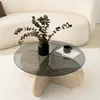 A round glass coffee table with a travertine base, decorated with a black vase of dried eucalyptus branches and a small patterned mug on a book. The table is placed on a white rug in front of a textured white sofa. Produktbild-Vorschau
