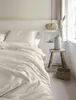 A neatly made bed with a textured duvet cover, a pillow, and a bedside table with a vase and a basket. Produktbild-Vorschau