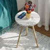 A small round side table with a marble-like top and wooden legs, placed on a rug next to a teal blanket. A smartphone and a vase of colorful flowers are on the table. Produktbild-Vorschau