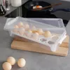 An egg holder filled with eggs sits on a wooden cutting board next to a frying pan with a fried egg. Podgląd zdjęcia produktu