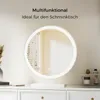 A round LED makeup mirror is displayed on a white dresser, with decorative items and makeup products nearby. The background is a softly lit room. Voir les images du produit