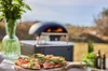 A pizza oven on a table with a pizza in the foreground, set in an outdoor garden setting. Produktbild-Vorschau