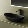 A black ceramic vessel sink with a black faucet is placed on a light green vanity with a fluted front. A lit mirror is visible in the background. Produktbild-Vorschau
