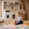 A toddler plays with toys in front of a wooden play kitchen. Produktbild-Vorschau