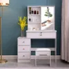 A white vanity table with a sliding mirror, drawers, and a stool, set against a teal wall. A floor lamp and a vase of yellow flowers are also visible. Voir les images du produit