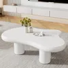 A white, irregularly shaped coffee table with two cylindrical legs, placed on a light gray rug in a living room setting. A vase with green foliage and a small decorative tray with a candle are on the table. Produktbild-Vorschau