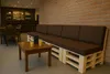 A long sofa made of pallets with brown cushions is placed against a brick wall in a cafe or lounge area. A small wooden table with two glasses of orange juice is in front of the sofa. Produktbild-Vorschau