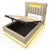 A beige upholstered bed frame with its mattress support lifted, revealing storage space underneath. The headboard and base have integrated yellow LED lighting. Náhled obrázku produktu