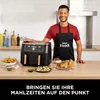 A man in an apron stands in a kitchen next to a dual-zone air fryer, with prepared food on the counter. Produktbild-Vorschau