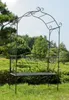 A black metal garden bench with an archway, set in a lush garden with green bushes and trees. Produktbild-Vorschau