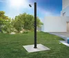 An outdoor shower is set up on a grassy lawn in front of a modern white house, with trees and rocks in the background. Produktbild-Vorschau