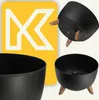 A collage of images showing a black plastic plant pot with wooden legs, and a yellow graphic with a white 'K' logo. Produktbild-Vorschau