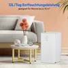 A white dehumidifier is placed on the floor next to a small round coffee table with decorative items and a sofa in the background. Produktbild-Vorschau