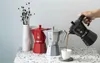 A hand is pouring coffee from a black Moka pot into a white cup, with a red and a silver Moka pot and a vase of dried flowers in the background. Produktbild-Vorschau