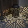 A black solar lantern with a leaf pattern casts intricate shadows on a textured surface and a wooden wall. Produktbild-Vorschau