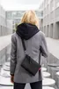 A person wearing a gray blazer and a black hoodie stands outdoors in front of a modern building, with a black crossbody bag. Produktbild-Vorschau