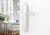 A close-up of a white door handle on a white wall, with a blurred background of a dining area. Produktbild-Vorschau