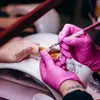 A close-up shot of a nail technician applying gel to a client's fingernail using a nail form and a brush. The technician is wearing pink gloves. Náhľad obrázku produktu