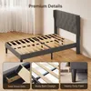 A dark gray upholstered bed frame with a wingback headboard is shown in a room with wooden floors and white curtains. Insets highlight the solid wood slats, mute bars design, and heavy-duty feet. Náhled obrázku produktu