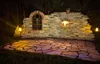 An outdoor patio area at night, featuring a rustic brick wall with a decorative arched window and a small lit alcove. The patio is paved with irregular flagstones, and illuminated by decorative garden lights. Produktbild-Vorschau