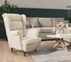 A cream-colored wingback armchair and a light-colored sofa in a living room setting with a coffee table and flowers.