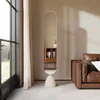 A modern, oval floor mirror with a beige base stands in a living room next to a brown leather sofa and a large window with sheer curtains. Produktbild-Vorschau