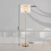 A floor lamp with a ribbed shade and a gold-colored stem and base, shown against a blurred background of furniture. Náhľad obrázku produktu