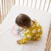 Baby in gelbem Strampler mit Wolkenmuster spielt mit rosa und blauem Lenkradspielzeug in einem Holzbettchen Náhľad obrázku produktu